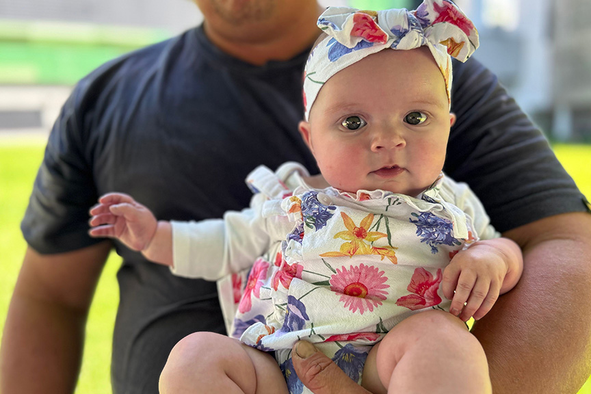 Ruby aged six months old with blue eyes wearing a white, pink, orange and blue floral bloomer and matching headband