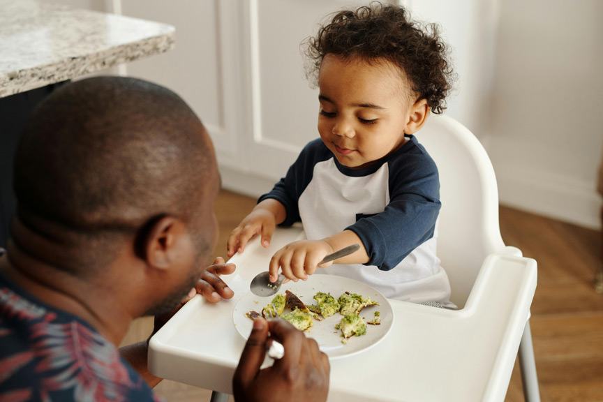 A toodler in high chair refusing to eat some food and her dad traying to feed her.