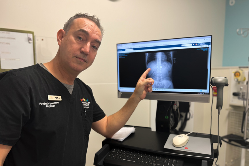 QCH paediatric emergency doctor, a middle aged man wearing black scrubs, points to an x ray of a round foreign object in child's abdomen.